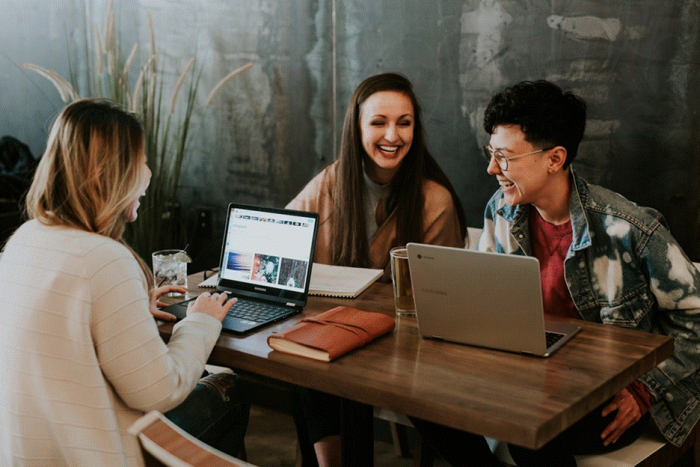 Title: Three colleagues talking and laughing around laptops at a shared table - Description: Three colleagues talking and laughing around laptops at a shared table