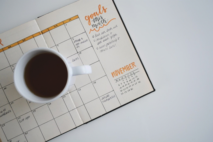 Title: Paper planner open to a monthly calendar next to a cup of coffee - Description: Paper planner open to a monthly calendar next to a cup of coffee
