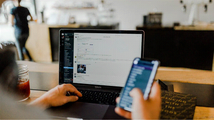 Title: Person scrolling a social media feed on a smartphone at a cafe table with an open laptop - Description: Person scrolling a social media feed on a smartphone at a cafe table with an open laptop