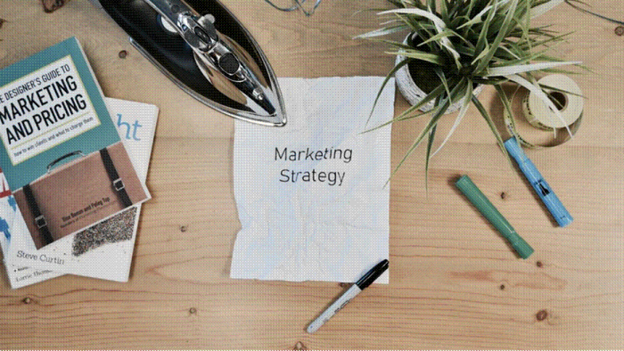 Title: Desk flat lay with a handwritten marketing strategy note, marketing books, pens and a small plant - Description: Desk flat lay with a handwritten marketing strategy note, marketing books, pens and a small plant