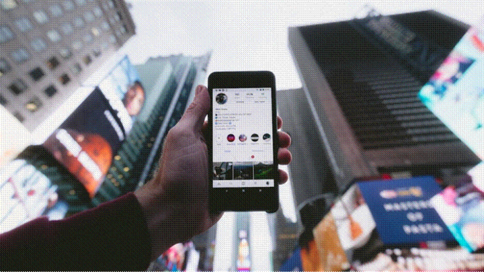 Title: Hand holding a smartphone showing an Instagram profile against a bright, busy city street - Description: Hand holding a smartphone showing an Instagram profile against a bright, busy city street