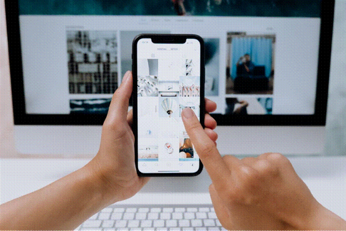 Title: Hands holding a smartphone while browsing a social feed in front of a desktop screen - Description: Hands holding a smartphone while browsing a social feed in front of a desktop screen