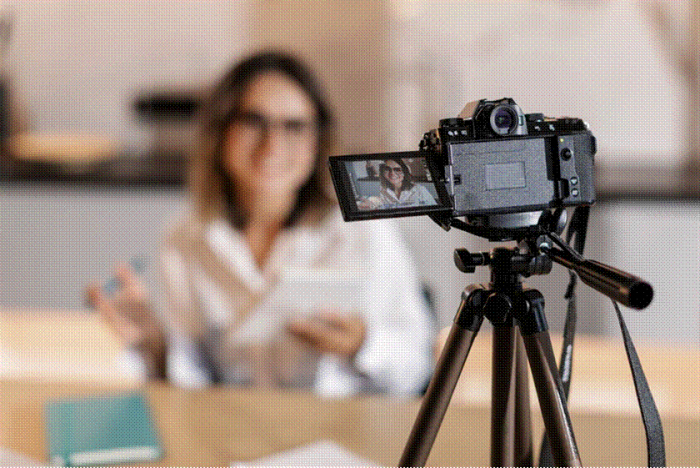 Title: A vlogger records to a camera mounted on a tripod at a home desk - Description: A vlogger records to a camera mounted on a tripod at a home desk