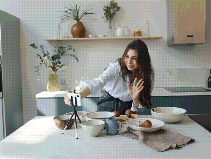 Title: A creator in a kitchen records a video on a smartphone mounted on a small tripod - Description: A creator in a kitchen records a video on a smartphone mounted on a small tripod
