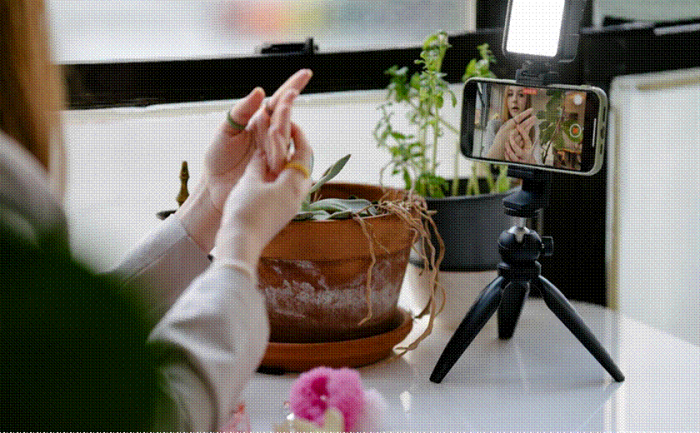 Title: A hand gestures toward a smartphone on a mini tripod with a small light, recording a take - Description: A hand gestures toward a smartphone on a mini tripod with a small light, recording a take