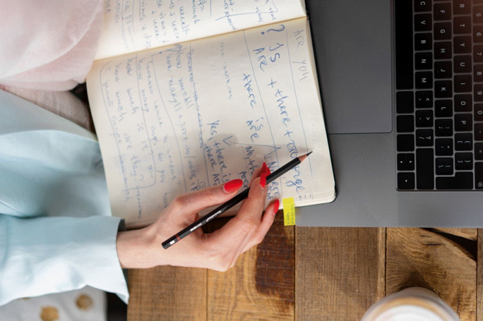Title: Handwritten bio drafts in a notebook beside a laptop - Description: Handwritten bio drafts in a notebook beside a laptop