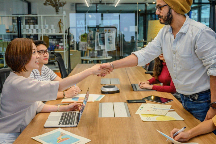 Title: Two people shaking hands across a table at a work meeting - Description: Two people shaking hands across a table at a work meeting