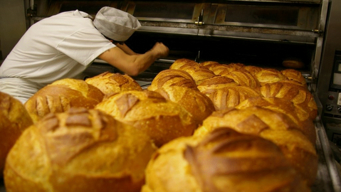 Title: Baker in whites loading an oven beside a rack of fresh crusty bread - Description: Baker in whites loading an oven beside a rack of fresh crusty bread