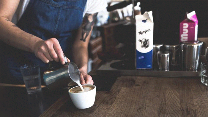 Title: Barista pouring steamed milk into a latte on a wooden cafe counter - Description: Barista pouring steamed milk into a latte on a wooden cafe counter